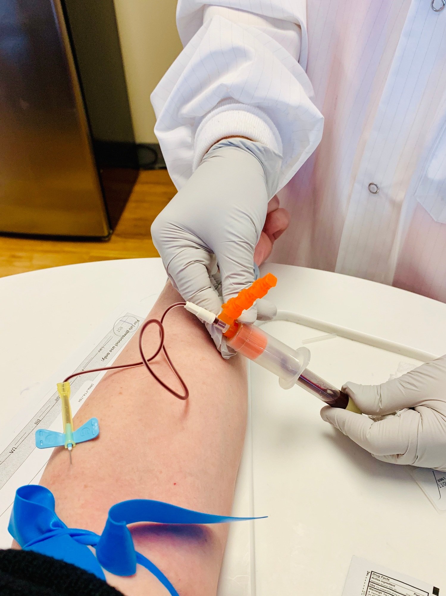 Phlebotomist wearing gloves is drawing blood from arm that has a tourniquet in place.